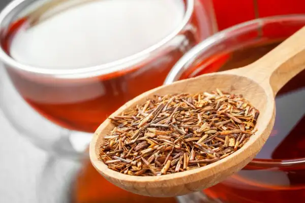 Rooibos Tee: Wirkung Schlafen. Eine Holzschaufel voller Rooibos-Blätter neben einer Tasse des roten Tees.