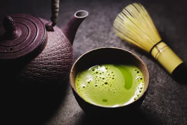 Eine traditionelle japanische Teekanne und ein Chasen (Bambusbesen) neben einer Schale mit schaumigem, leuchtend grünem Matcha. So wird Matcha zubereitet.