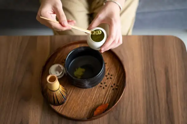 Vorbereitung von Matcha für die Schwangerschaft: Hände schöpfen grünes Pulver in eine Schale mit heißem Wasser. Ein Chasen (Bambusbesen) liegt daneben.